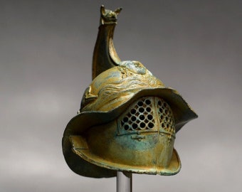 Peut inclure: Un casque de gladiateur en bronze avec une patine verte, présenté sur un support en acrylique transparent. Le casque a une crête distinctive et un masque facial avec un motif en grille.