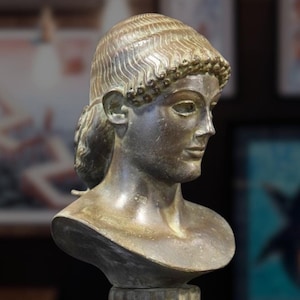 Könnte beinhalten: Bronzebüste einer Frau mit geflochtener Frisur, die eine Krone trägt. Die Büste steht auf einem Sockel mit einem geriffelten Sockel.