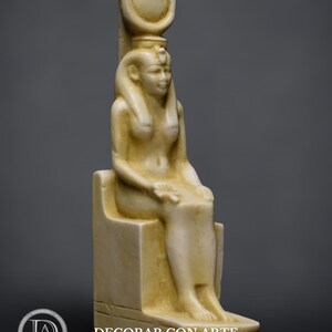 Puede incluir: Una escultura de piedra blanca de una diosa egipcia sentada en un trono. La diosa lleva una corona y una larga t&uacute;nica fluida. La escultura mide aproximadamente 25 cm de alto.