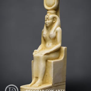 Puede incluir: Una estatua de m&aacute;rmol blanco de una diosa egipcia sentada en un trono. Lleva un alto tocado con un disco solar y una serpiente uraeus. La estatua es detallada y tiene un acabado liso.