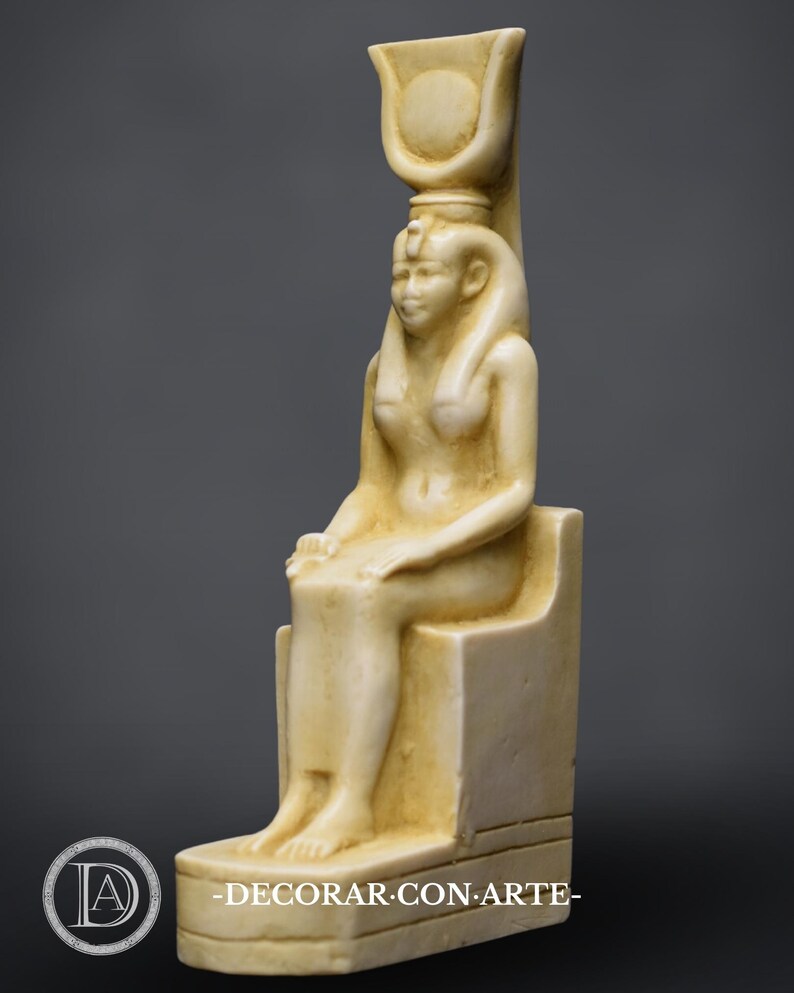 Puede incluir: Una estatua de m&aacute;rmol blanco de una diosa egipcia sentada en un trono. Lleva un alto tocado con un disco solar y una serpiente uraeus. La estatua es detallada y tiene un acabado liso.