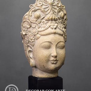 May include: A beige ceramic sculpture of a Buddha head with closed eyes, wearing a detailed crown with floral motifs. The sculpture is mounted on a black pedestal with the text "DECORAR.CON.ARTE- ".