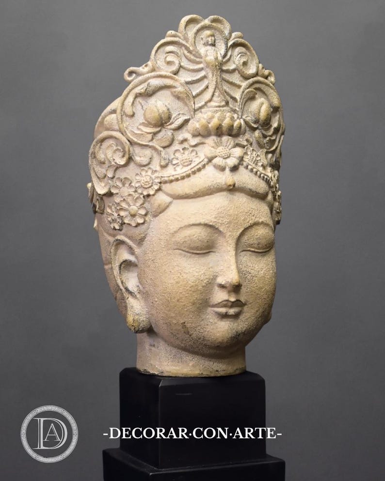 May include: A beige ceramic sculpture of a Buddha head with closed eyes, wearing a detailed crown with floral motifs. The sculpture is mounted on a black pedestal with the text "DECORAR.CON.ARTE- ".