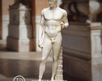 May include: A classical white marble statue of a nude male figure. The statue depicts a young man with a cloth wrapped around his head, standing on a textured base. The statue is in a classical style, with detailed musculature and a serene expression.