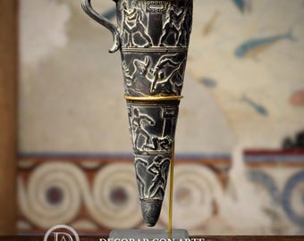 May include: A dark gray, cone-shaped decorative cup with a handle and gold band. The cup features detailed relief carvings of figures in action. The cup is displayed on a gray rectangular base with the text "-DECORAR CON ARTE-" at the bottom.