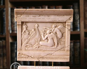 May include: A rectangular beige stone relief sculpture featuring a carved scene of a boat with figures and winged beings playing a lyre. The top section contains Greek text and the base is supported by two thin metal rods on a stone stand.