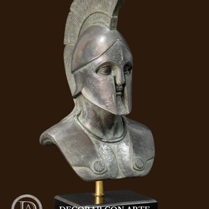 May include: A bronze-colored bust of a warrior wearing a helmet with a tall crest. The bust is mounted on a black rectangular base. The warrior's armor includes shoulder decorations. The bust is a decorative item.