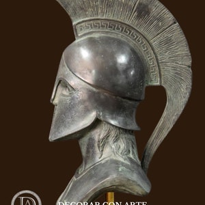 May include: A bronze-colored bust of a Spartan warrior, featuring a detailed helmet with a crest and a decorative border. The bust is mounted on a gold-colored stand, with the text "DECORAR.CON.ARTE" visible.