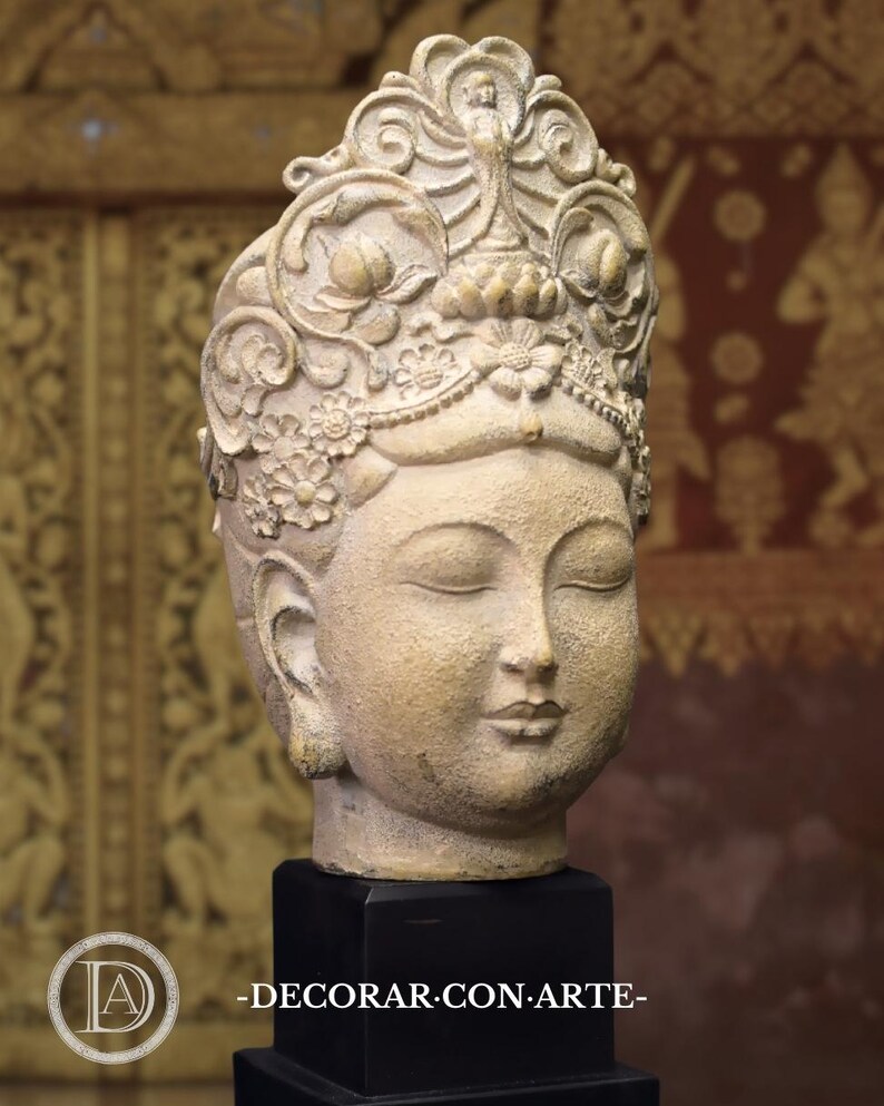 May include: A beige stone sculpture of a woman's head with a detailed crown and closed eyes. The sculpture is on a black base.