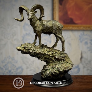 Peut inclure: Une sculpture de bélier de couleur bronze avec de grands cornes incurvées se dresse sur une base rocheuse texturée. La sculpture est montée sur une base circulaire noire. Le texte "-DECORAR CON ARTE" est visible en bas.