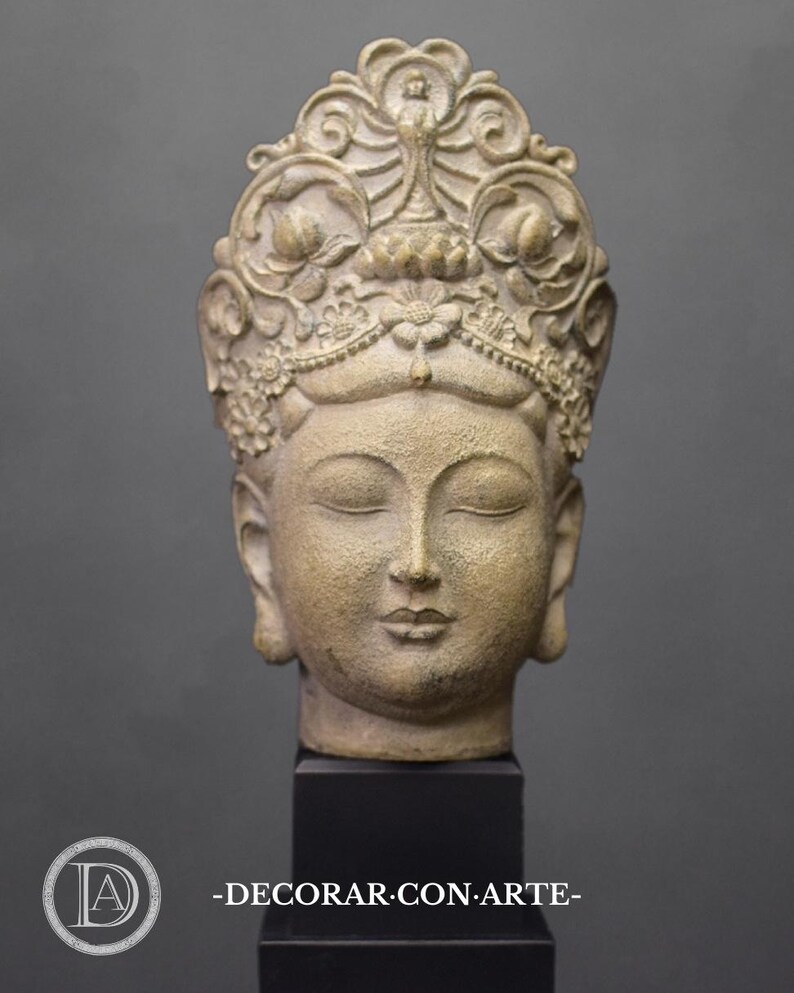 May include: A beige stone sculpture of a Buddha head with closed eyes and a crown of intricate floral designs. The sculpture is mounted on a black base.