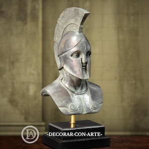 May include: A bronze-toned bust of a classical warrior, complete with a helmet and chest armor. The sculpture is mounted on a black rectangular base. The text "-DECORAR-CON-ARTE-" is visible on the base.