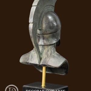 May include: Bronze-colored bust of a classical warrior, featuring a detailed helmet with a prominent crest. The sculpture is mounted on a black rectangular base with a gold-colored support rod. The bust is a decorative item.
