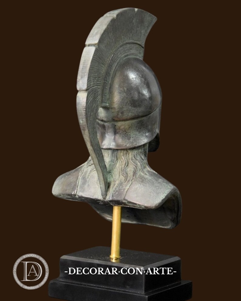 May include: Bronze-colored bust of a classical warrior, featuring a detailed helmet with a prominent crest. The sculpture is mounted on a black rectangular base with a gold-colored support rod. The bust is a decorative item.
