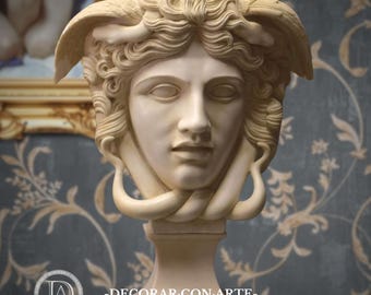Op de afbeelding: Een crèmekleurige Medusa-hoofdsculptuur op een voetstuk. De sculptuur heeft gedetailleerde gelaatstrekken, vloeiend haar en slangachtige elementen. De achtergrond omvat een ingelijst schilderij en een behang met patronen. De tekst "-DECORAR CON ARTE-" is zichtbaar.