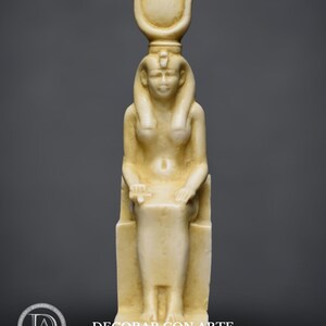 Puede incluir: Una estatua de cer&aacute;mica blanca de una diosa egipcia sentada en un trono. La diosa lleva un alto tocado con un disco solar y una serpiente uraeus. Tiene una expresi&oacute;n serena y sostiene un bast&oacute;n en su mano derecha.