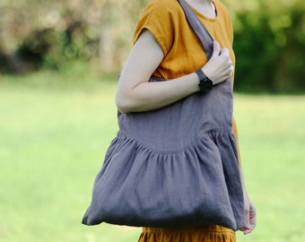 May include: A gray linen tote bag with a ruffled bottom. The bag is being held by a person wearing a yellow dress.