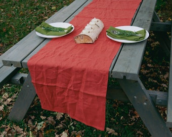 May include: A red linen table runner with a natural wood log centerpiece and two white plates with green napkins on a gray wooden picnic table.