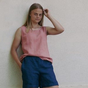 May include: A woman wearing a sleeveless, dusty rose linen tank top and dark blue linen shorts. She is standing in front of a white wall.