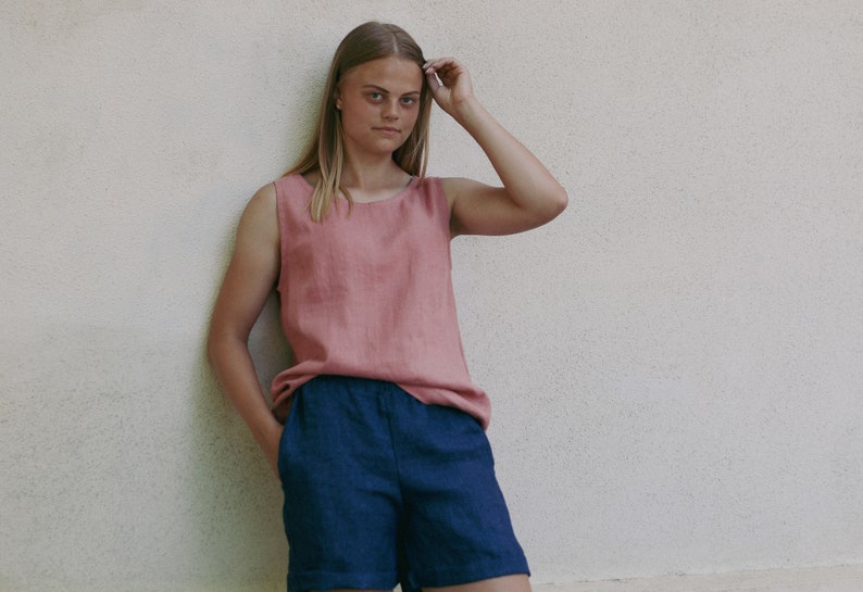 May include: A woman wearing a sleeveless, dusty rose linen tank top and dark blue linen shorts. She is standing in front of a white wall.