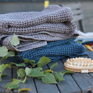 May include: A stack of two waffle weave towels, one grey and one blue, with a wooden brush with natural bristles on a wooden table with green leaves.