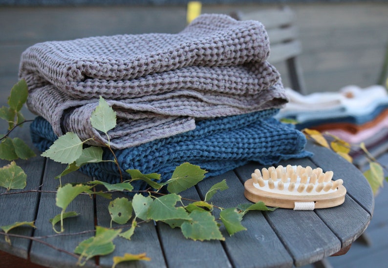 May include: A stack of two waffle weave towels, one grey and one blue, with a wooden brush with natural bristles on a wooden table with green leaves.