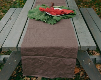 May include: A brown linen table runner with a red napkin and a plate with red apples on a green leaf on a wooden picnic table.