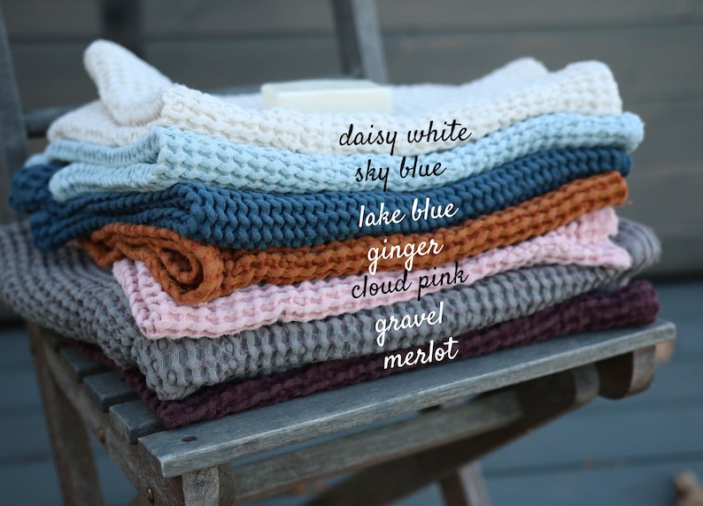 May include: A stack of waffle weave cotton throws in various colours, including daisy white, sky blue, lake blue, ginger, cloud pink, gravel, and merlot.