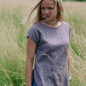 May include: A woman wearing a loose-fitting, grey linen top with short sleeves. She is standing in a field of tall grass.