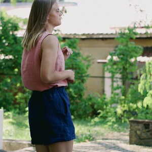 May include: A woman wearing a pink sleeveless top and dark blue linen shorts. She is standing outdoors with greenery in the background.