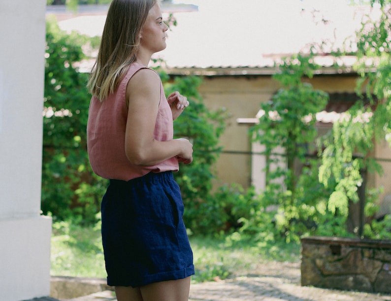 May include: A woman wearing a pink sleeveless top and dark blue linen shorts. She is standing outdoors with greenery in the background.