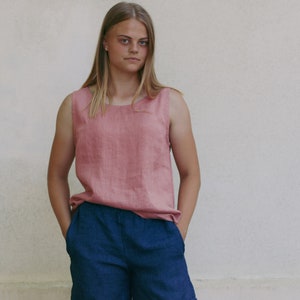 May include: A woman wearing a sleeveless, pink linen top and dark blue shorts. She is standing in front of a white wall.