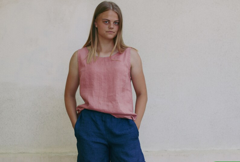 May include: A woman wearing a sleeveless, pink linen top and dark blue shorts. She is standing in front of a white wall.
