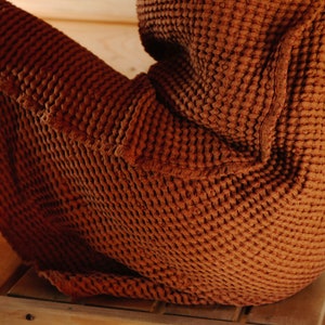 May include: A brown waffle-weave robe with a pocket on the side. The robe is worn by a person sitting on a wooden bench.