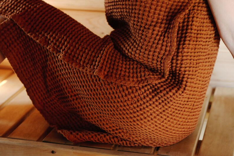 May include: A brown waffle-weave robe with a pocket on the side. The robe is worn by a person sitting on a wooden bench.