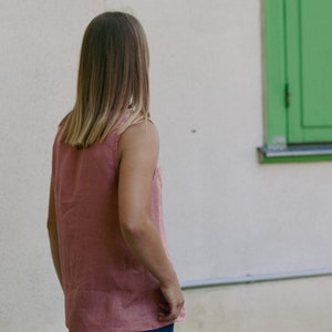 May include: A woman wearing a sleeveless, pink top with her back to the camera. She is standing in front of a white wall with a green window.