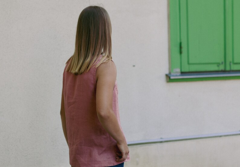May include: A woman wearing a sleeveless, pink top with her back to the camera. She is standing in front of a white wall with a green window.