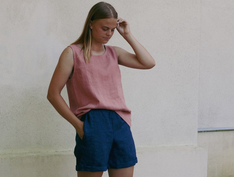 May include: A woman wearing a sleeveless, dusty rose linen top and dark blue linen shorts. The top has a scoop neck and the shorts have a cuffed hem.