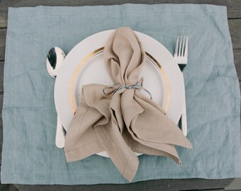 May include: A white plate with a gold rim and a beige linen napkin tied with string. The plate is on a blue linen placemat.