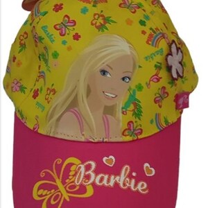 Könnte beinhalten: Eine gelbe Baseballkappe mit rosafarbenem Schirm. Die Kappe zeigt ein farbenfrohes Schmetterlingsdesign, einen Regenbogen und ein Bild von Barbie. Der Schirm hat einen rosafarbenen Schmetterling und das Wort "Barbie" in Weiß.