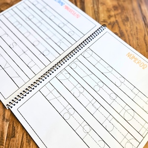 May include: A spiral-bound educational workbook featuring white pages with black dashed lines for tracing lowercase letters and numbers. The left page displays the alphabet, while the right page shows the text N&Uacute;MEROS and rows of numbers for practice.