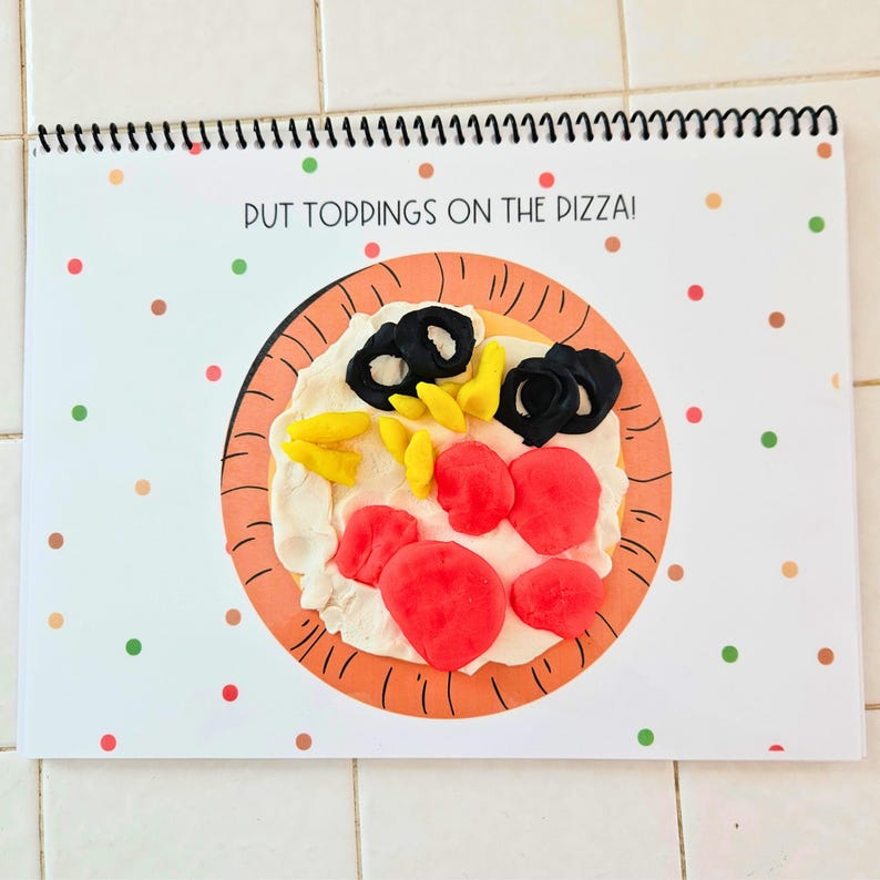May include: A white paper booklet with a colorful play dough pizza on the front. The pizza has red tomato slices, yellow corn, and black olives. The text on the front of the booklet says "Put toppings on the pizza!"