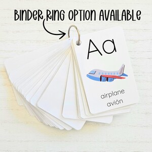 May include: A set of alphabet flash cards with a picture of an airplane and the letter "Aa" on the front. The words "airplane" and "avi&oacute;n" are written below the picture. The cards are arranged in a fan shape and have a metal ring attached to the top.