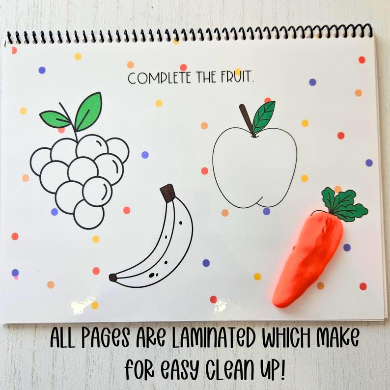 May include: A white, laminated activity book with colorful dots on the cover. The book has a spiral binding and the text "COMPLETE THE FRUIT." is printed on the cover. The book includes outlines of a banana, apple, grapes, and a carrot.