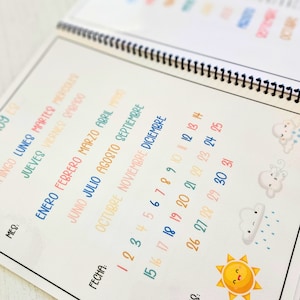 May include: A spiral-bound planner page featuring Spanish text for days of the week and months of the year in colorful, handwritten-style fonts. The page includes a numbered calendar from 1 to 31, with cute weather-themed illustrations of clouds and a sun.
