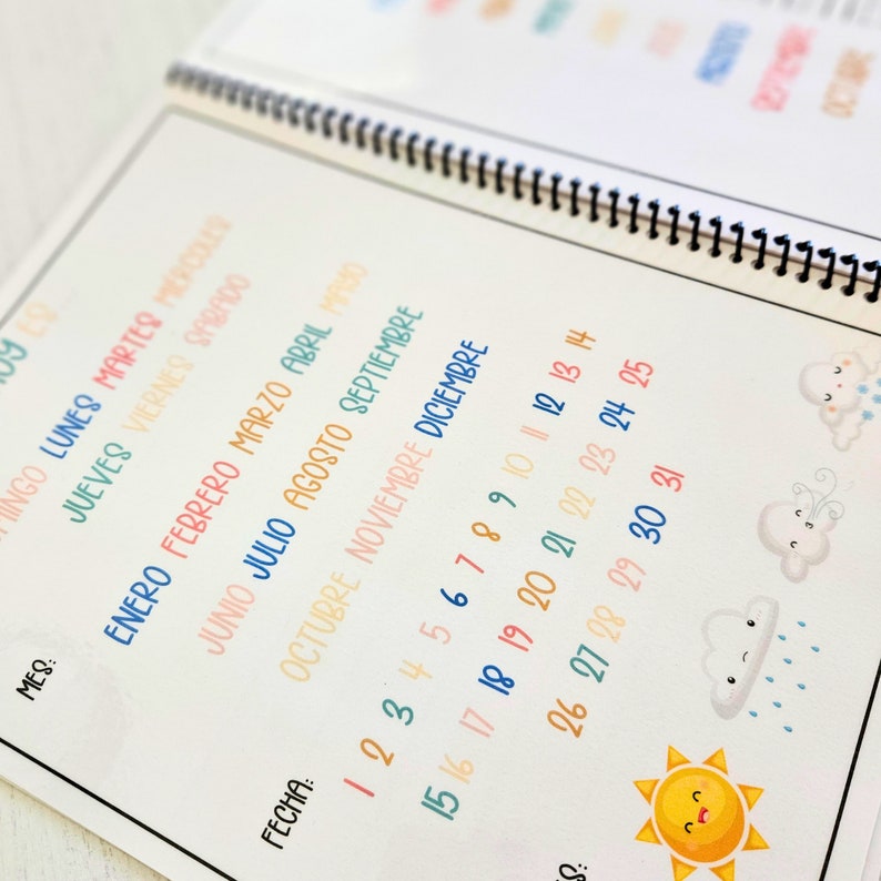 May include: A spiral-bound planner page featuring Spanish text for days of the week and months of the year in colorful, handwritten-style fonts. The page includes a numbered calendar from 1 to 31, with cute weather-themed illustrations of clouds and a sun.
