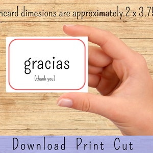 May include: A white flashcard with a red border, featuring the Spanish word "gracias" in black text, with the English translation "(thank you)" in smaller text below. The flashcard is being held by a hand against a woodgrain background.