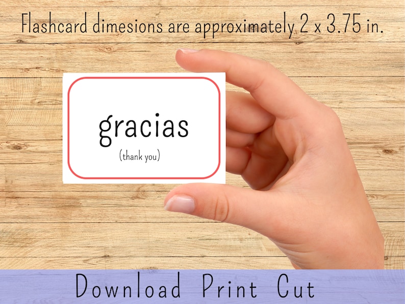 May include: A white flashcard with a red border, featuring the Spanish word "gracias" in black text, with the English translation "(thank you)" in smaller text below. The flashcard is being held by a hand against a woodgrain background.