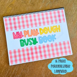 Puede incluir: Un libro de actividades con encuadernación en espiral y un borde de cuadros vichy rosa y blanco. La portada presenta el texto "MY PLAY DOUGH BUSY BOOK" en letras coloridas. Un gráfico azul en forma de nube muestra "16 PAGES PERSONALIZABLE LAMINATED".