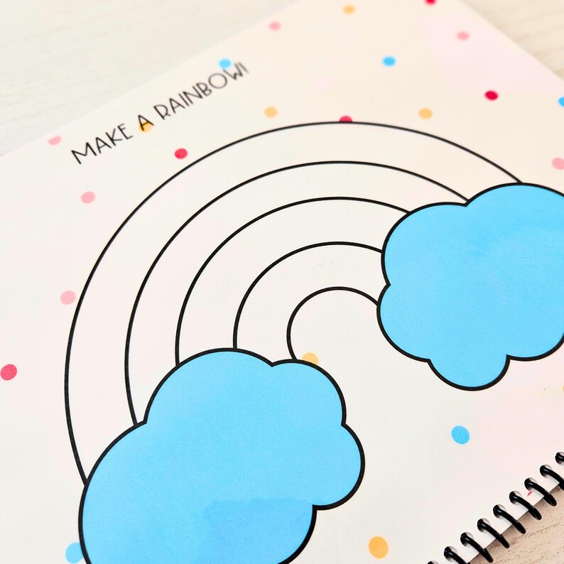 May include: A white coloring book with a black outline of a rainbow and clouds. The book has the words "Make a Rainbow" printed on the front cover. The background is a light pink with colorful dots.
